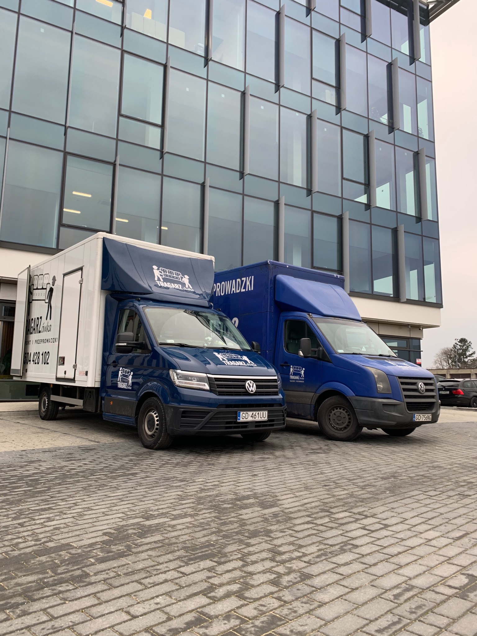 Dwa niebieskie samochody dostawcze firmy przeprowadzkowej 'Tragarzówka' zaparkowane na brukowanym parkingu przed nowoczesnym, przeszklonym biurowcem. Samochody mają białe i niebieskie nadwozia...