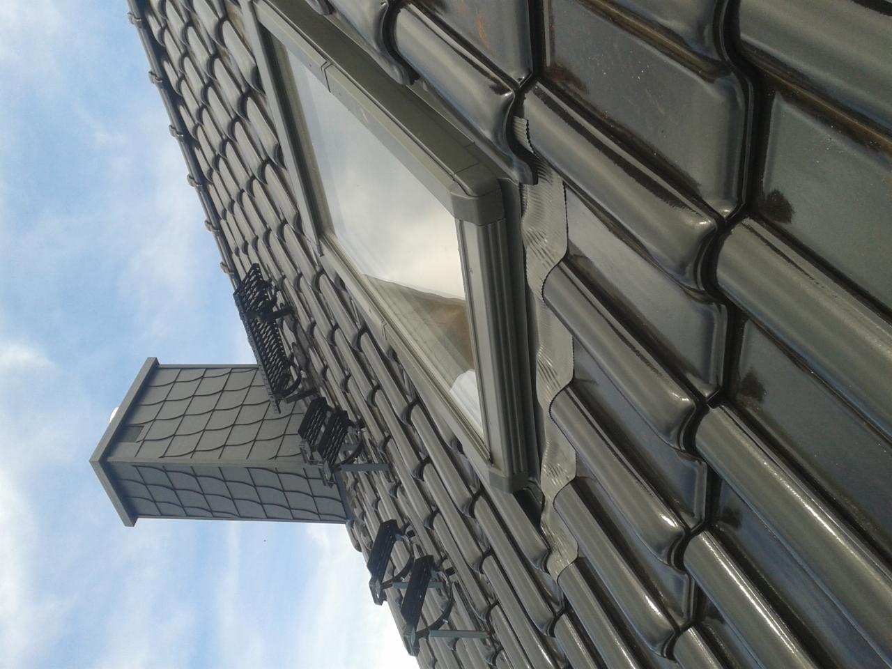 Błyszczące, grafitowe dachówki ceramiczne ułożone na dachu, widoczne okno dachowe z szarą ramą oraz fragment rynny i komina pokrytego dachówką karpiówką na tle błękitnego nieba.