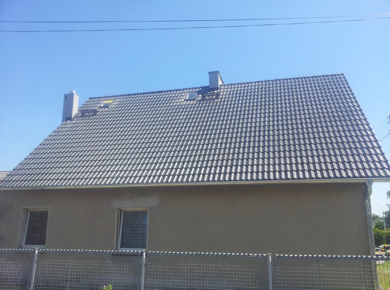 Dom jednorodzinny z szarą dachówką ceramiczną, widoczny komin, rynna i ogrodzenie siatkowe na tle błękitnego nieba.