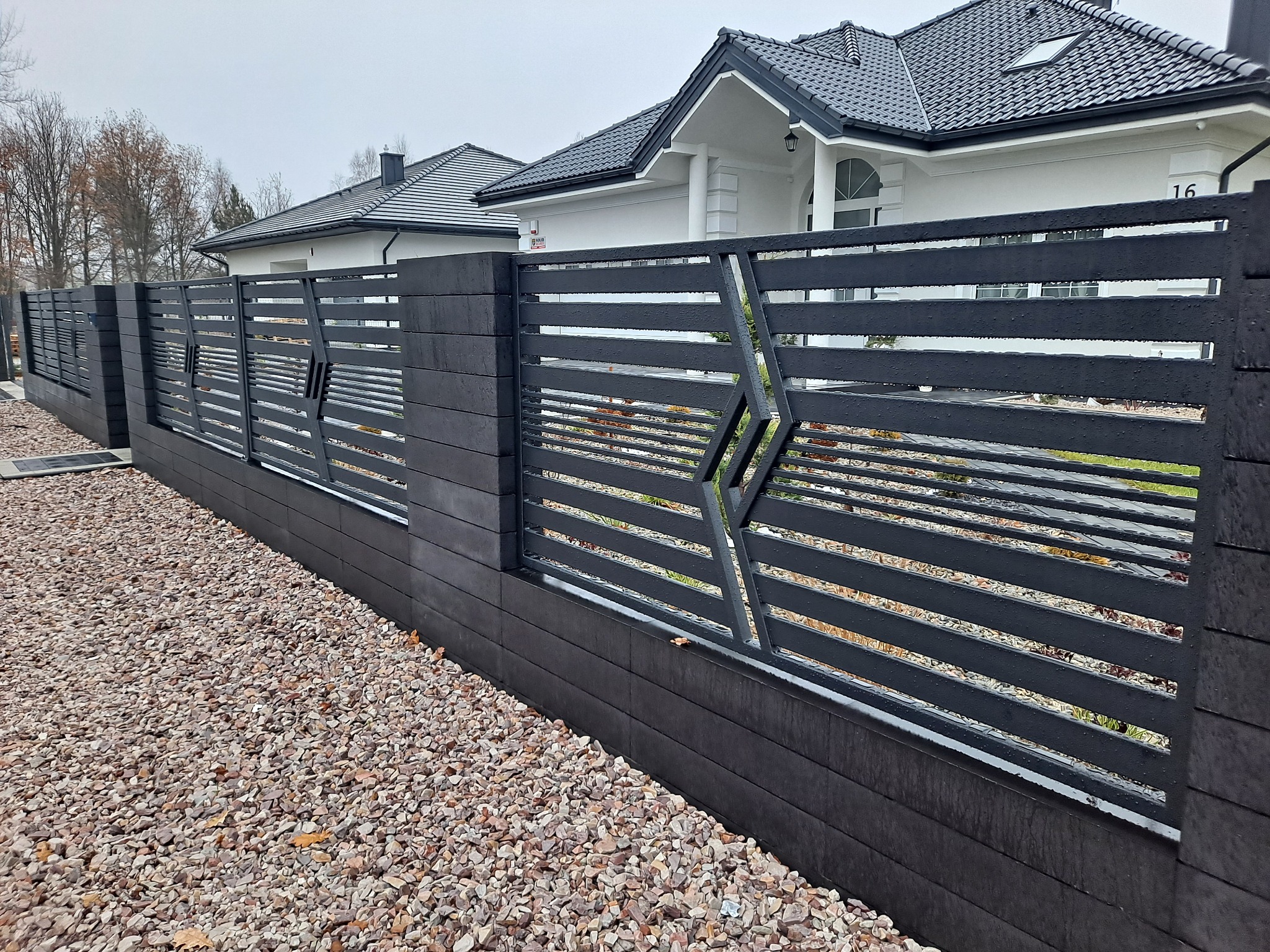 Nowoczesne, poziome ogrodzenie z metalowymi przęsłami w kolorze antracytowym, osadzone na niskiej podmurówce z imitacji drewna, z dekoracyjnymi elementami w kształcie rombów, na tle białego domu...