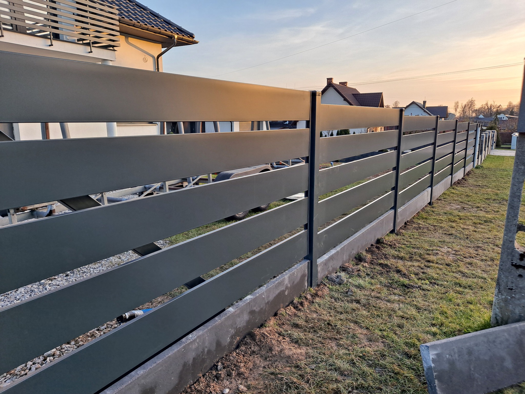 Nowoczesne ogrodzenie z poziomych, szarych paneli na betonowej podmurówce, widok z perspektywy trawnika w kierunku domu i zachodzącego słońca.