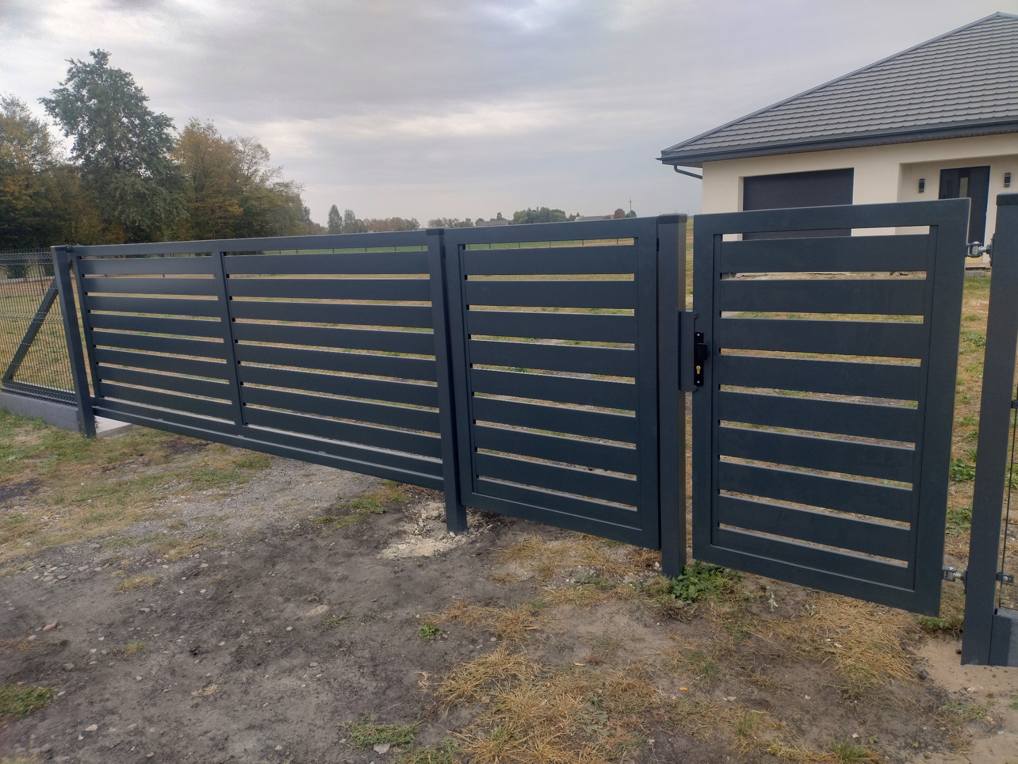 Nowoczesna, antracytowa brama wjazdowa z poziomymi panelami na tle domu z szarą dachówką i trawnika z widoczną siatką ogrodzeniową po lewej stronie.