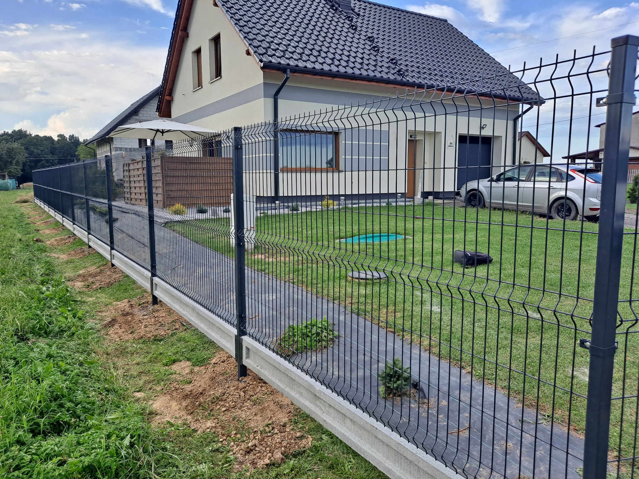 Długi, prosty odcinek czarnego ogrodzenia panelowego z podmurówką betonową, rozciągający się wzdłuż trawnika przed domem z szarą elewacją i ciemnym dachem, z widocznym fragmentem samochodu...