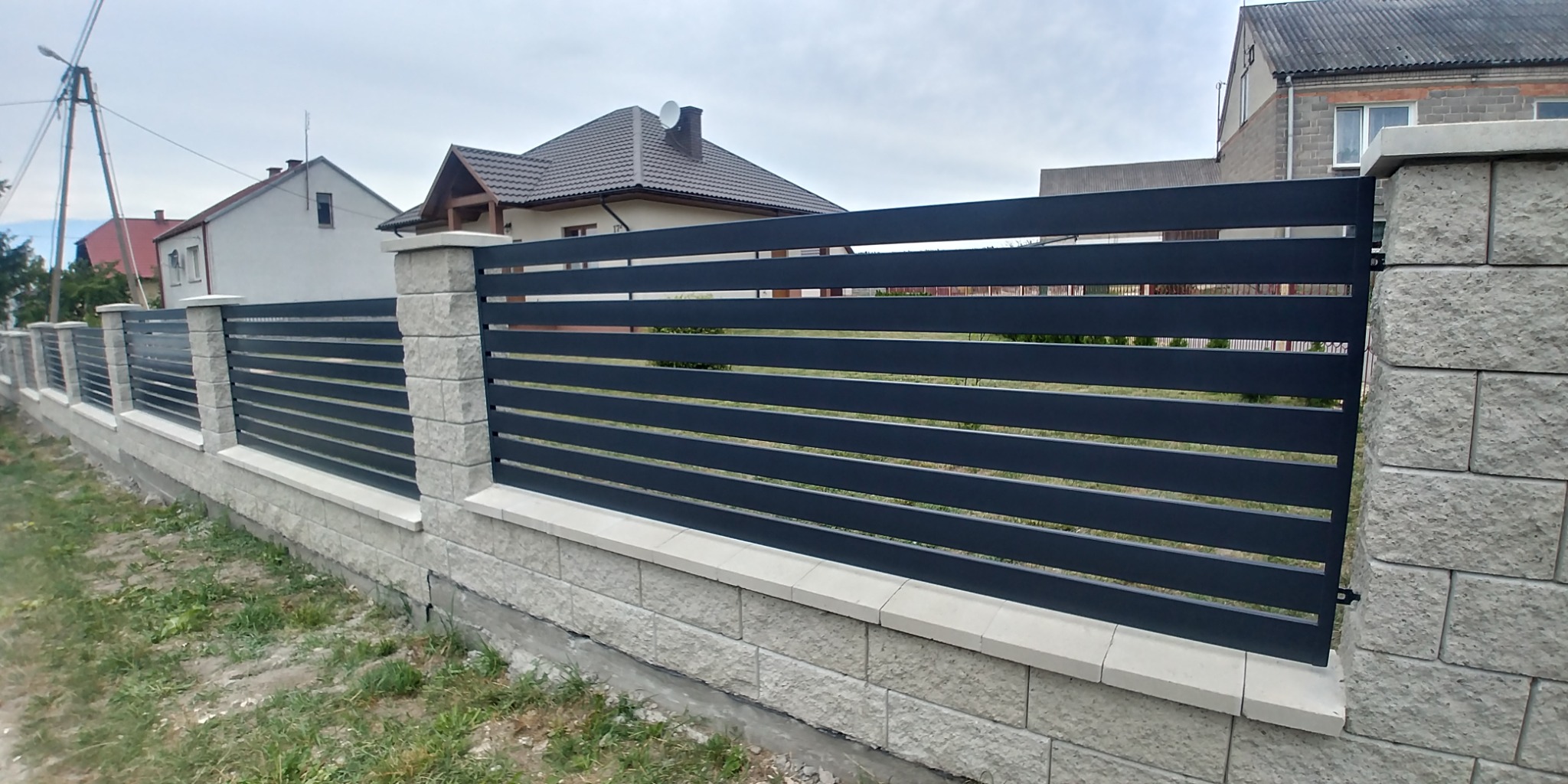 Nowoczesne ogrodzenie z poziomych, grafitowych paneli na niskiej podmurówce z szarej kostki, z widocznymi słupkami z tego samego materiału, na tle domów.