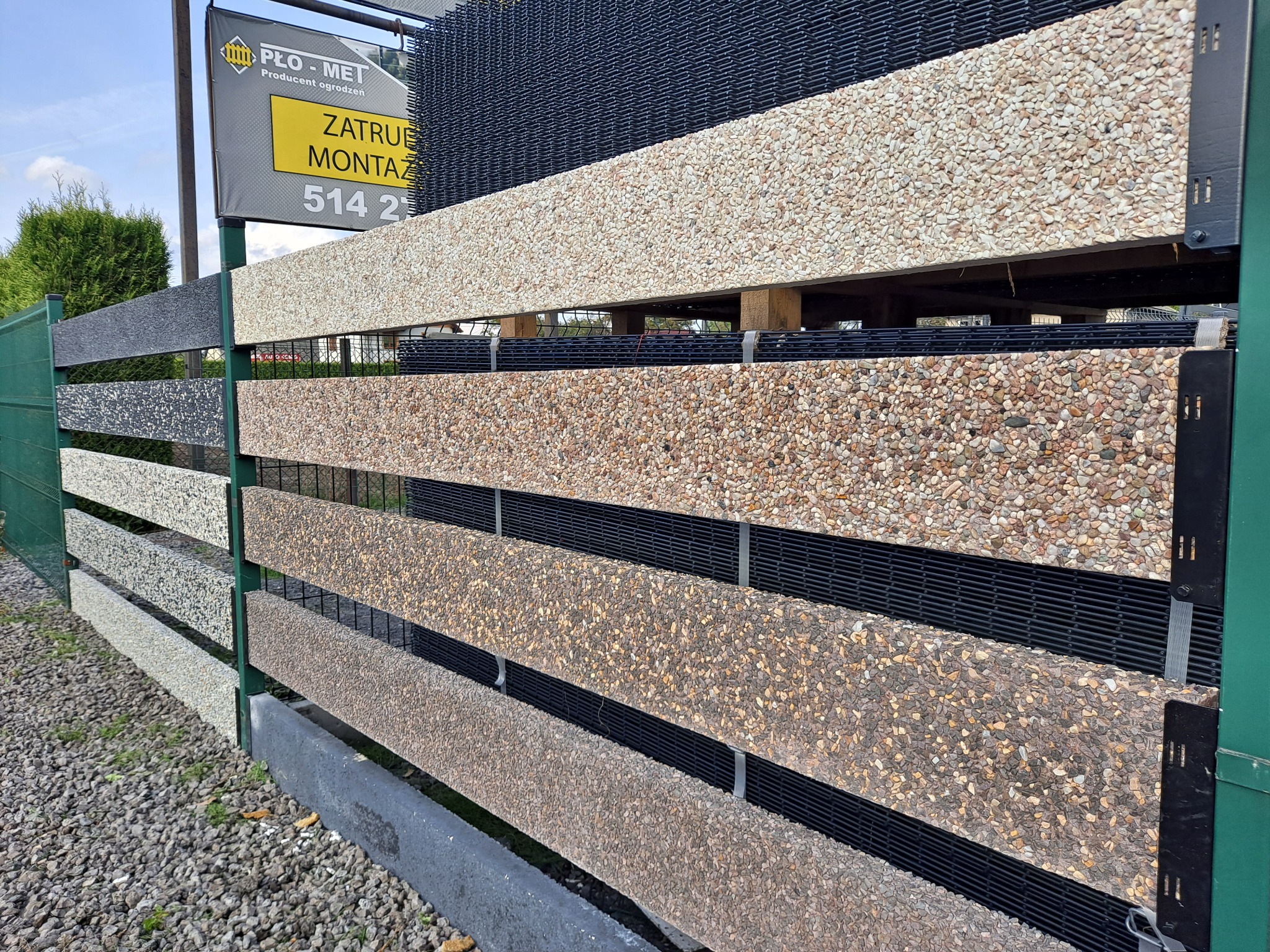Ekspozycja fragmentów ogrodzenia z elementami betonowymi i siatką, prezentacja różnych wariantów kolorystycznych i faktur kruszywa na tle żwirowego podłoża i tablicy reklamowej producenta.