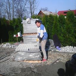 Sakazo - Mężczyzna w roboczym stroju przygotowuje kostkę brukową na palecie, otoczoną żwirem i zielenią, w tle dom z czerwonym dachem.