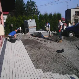 Układanie kostki granitowej: trzech pracowników w trakcie pracy nad podjazdem z kostki granitowej, widoczne ułożone fragmenty i palety z kostką.