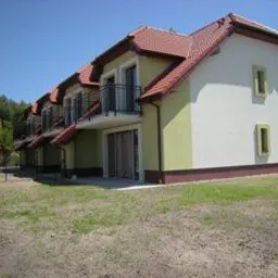 Dwukondygnacyjny budynek mieszkalny z balkonami, otynkowany na biało-żółto, z czerwoną dachówką, na tle zielonego trawnika i błękitnego nieba.