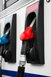 Dwa pistolety do tankowania paliwa na dystrybutorze: czerwony i niebieski, z metalowymi wylewkami i czarnymi obudowami, na tle białej ściany dystrybutora.