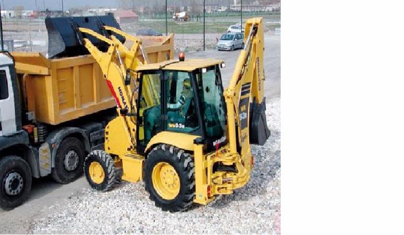 Żółta koparko-ładowarka Komatsu 93R wysypuje ziemię do żółtej ciężarówki na żwirowym podłożu, w tle widoczny krajobraz z drzewami i zaparkowanymi samochodami.