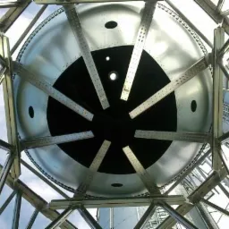 Wnętrze stalowej konstrukcji hali z widocznym metalowym zbiornikiem od dołu, oświetlonym naturalnym światłem. Widoczne elementy konstrukcyjne i fragment nieba.