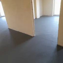 Świeżo wylana, gładka, szara posadzka jastrychowa w pomieszczeniu z surowymi ścianami działowymi i oknami, widoczne delikatne refleksy światła.