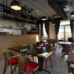 Jasne wnętrze restauracji z drewnianym barem, stolikami z nakryciami i czerwonymi krzesłami, oświetlone wiszącymi lampami.