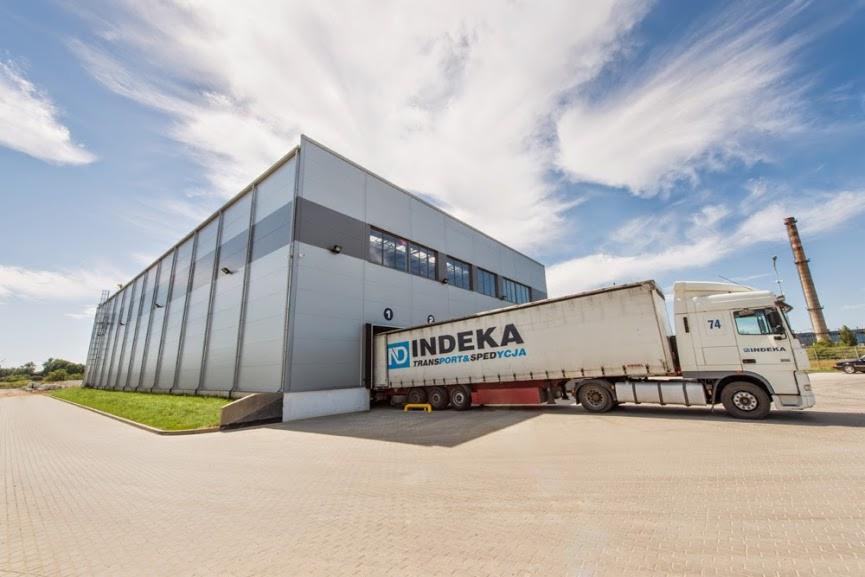 Szara hala magazynowa z rampą, do której podjeżdża biała ciężarówka z logo Indeka Transport i Spedycja, widok z zewnątrz w słoneczny dzień.