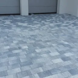 Świeżo ułożona, szara kostka brukowa przed garażem, widoczna łopata i stos kostki do wykończenia krawędzi.