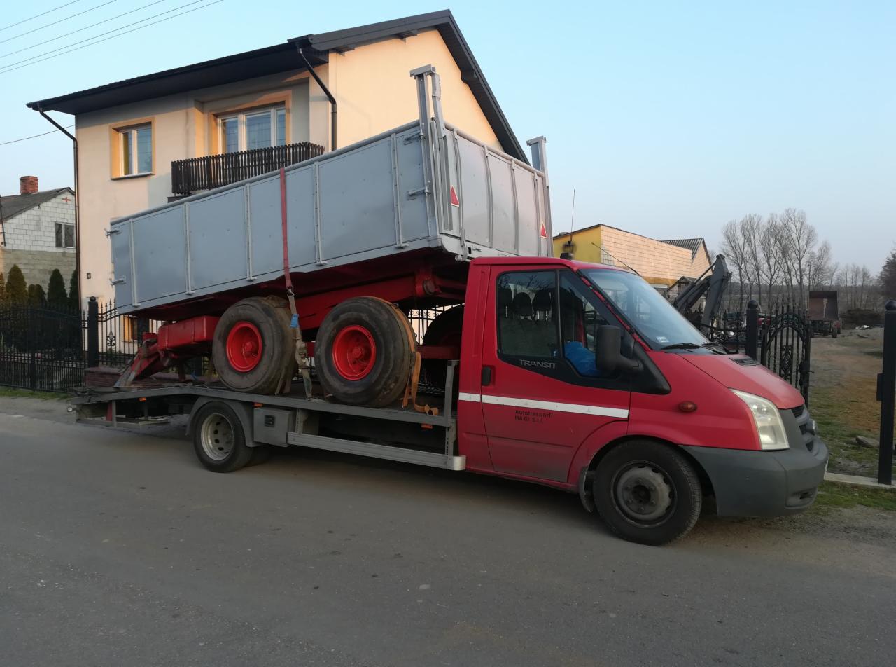 Czerwony samochód pomocy drogowej Ford Transit z platformą transportową przewozi szarą przyczepę rolniczą na tle zabudowań.