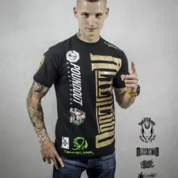 Młody mężczyzna z tatuażami prezentuje czarną koszulkę z dużym złotym nadrukiem POUNDOUT i logotypami sponsorów, wskazując palcem do góry, na tle szarej ściany.