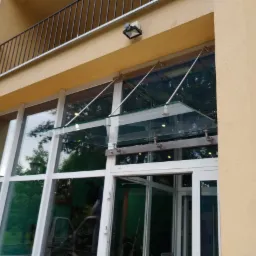 Szklany daszek nad wejściem do budynku z białymi ramami okiennymi i drzwiowymi, wsparty na metalowych wspornikach, widoczny z zewnątrz, z balkonem z czarną balustradą na piętrze.