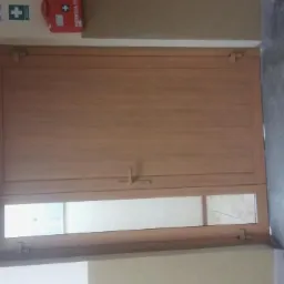 Zamknięte, dwuskrzydłowe drzwi z drewnopodobną okleiną i przeszkleniem w dolnej części, zamontowane wewnątrz budynku z widoczną apteczką i tabliczkami informacyjnymi na ścianie.