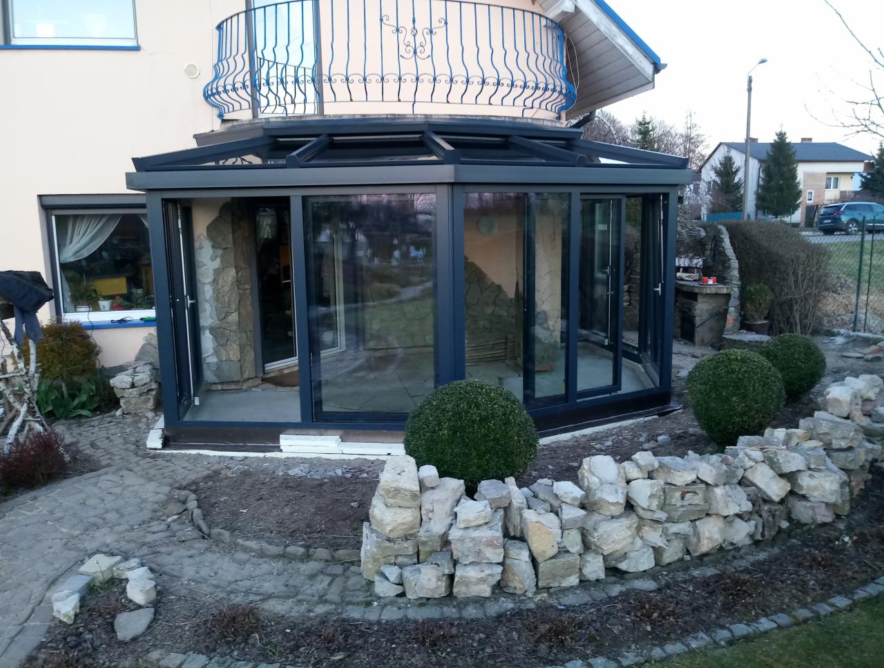 Szklana zabudowa tarasu z ciemną ramą, przylegająca do budynku z balkonem, otoczona kamiennym murkiem i kulistymi krzewami.