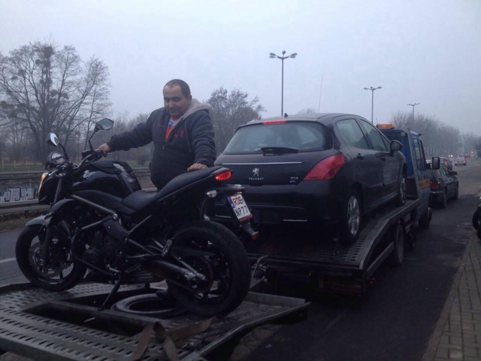 Czarny motocykl i Peugeot na lawecie. Mężczyzna w kurtce stoi obok. Transport lawetą na tle miejskiej ulicy w pochmurny dzień.