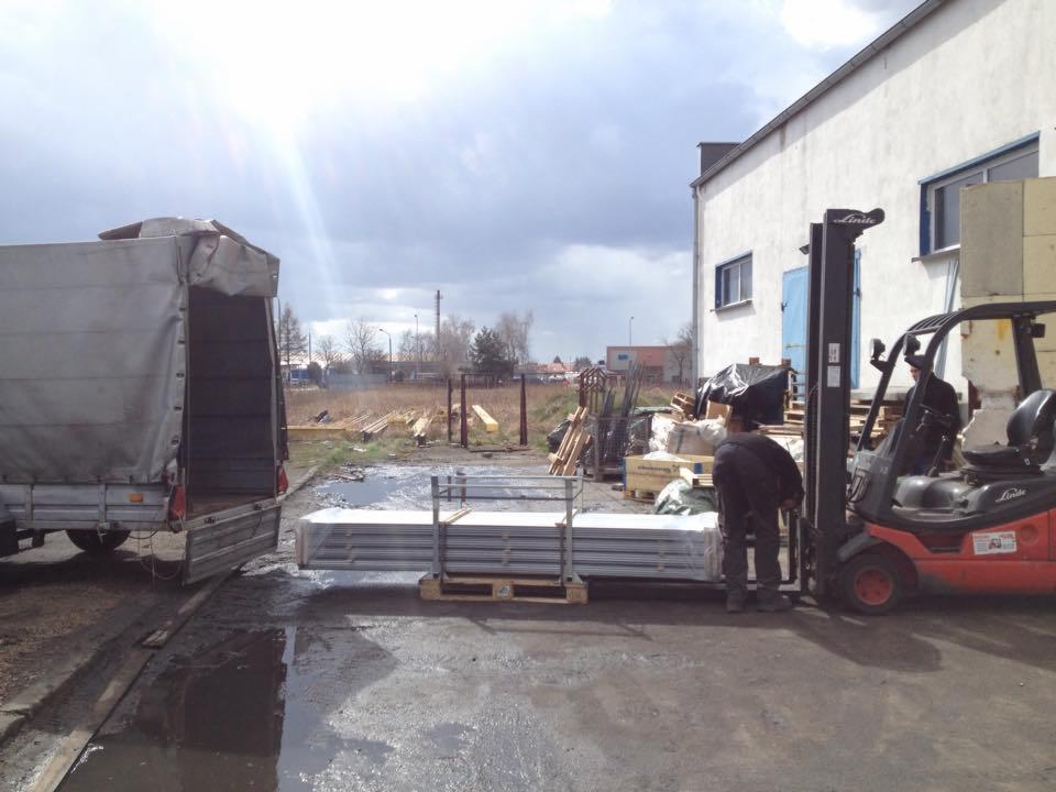 Wózek widłowy Linde załadowuje paletę z panelami do otwartej przyczepy busa na placu budowy; w tle budynek z niebieskimi oknami i pochmurne niebo.