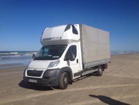 Biały bus dostawczy z plandeką marki Lamberet na piaszczystej plaży, z widocznym morzem i błękitnym niebem w tle. Nietypowa lokalizacja transportu.