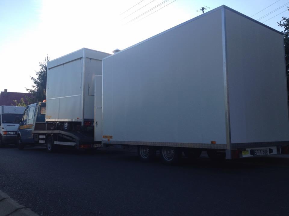 Laweta z dwoma kontenerami transportowymi na tle ulicy i zabudowań. Kontenery w kolorze białym, ustawione jeden za drugim. Widoczne tablice rejestracyjne.