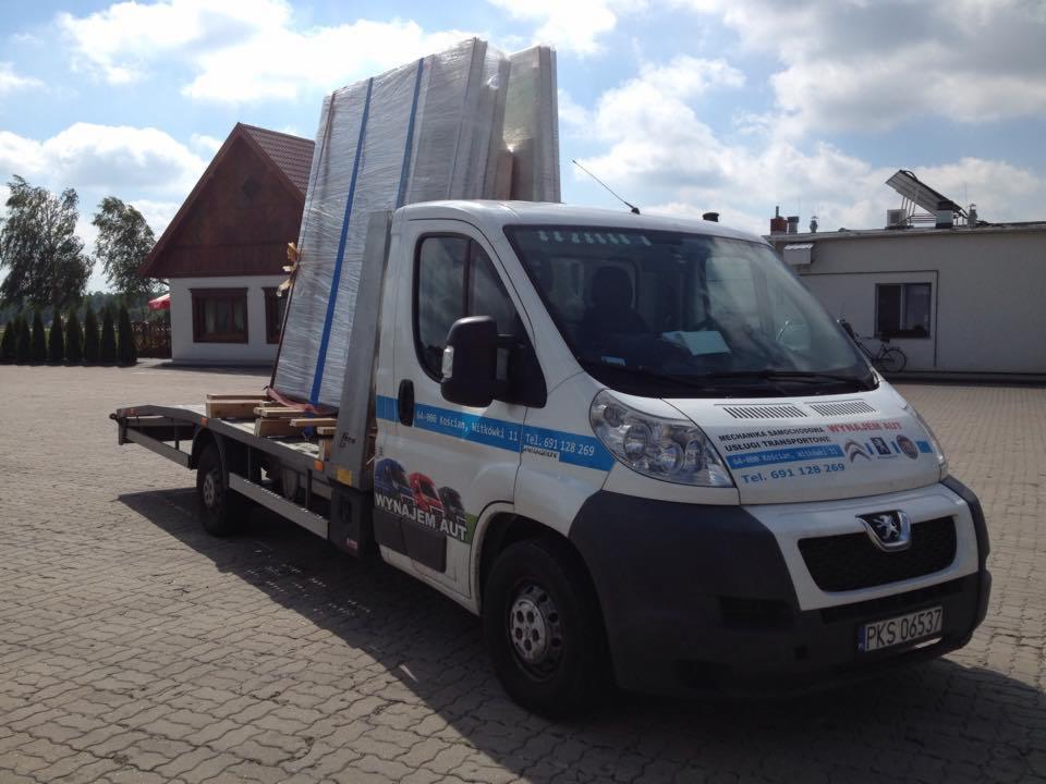 Biały bus z platformą transportową, przewożący zabezpieczone ładunki, na tle budynków i drzew, w słoneczny dzień. Widoczny numer rejestracyjny pojazdu.