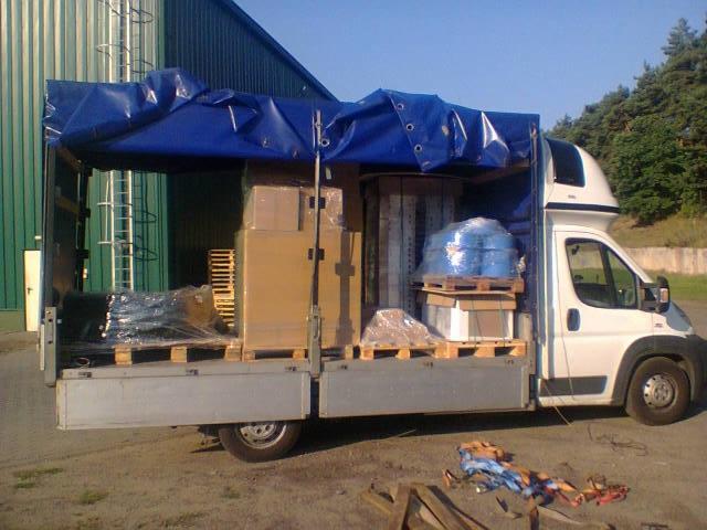 Otwarty bus transportowy z niebieską plandeką, załadowany kartonami i paletami z towarem. Widoczne elementy mocujące ładunek. Tło: budynek i drzewa.