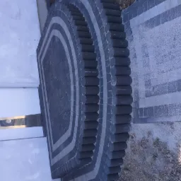 Półokrągła kompozycja z ciemnej kostki brukowej, obrzeża z walcowanych elementów, kontrastująca z jasną kostką na chodniku i elewacją budynku w tle.