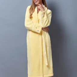 Uśmiechnięta blondynka w długim, żółtym, frotte szlafroku z kolorowymi kropkami na mankietach, pozuje na szarym tle.