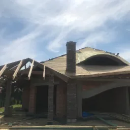 Budowa domu jednorodzinnego z nietypową konstrukcją dachu, widoczna więźba dachowa, komin z cegły klinkierowej i częściowo ułożone pokrycie dachowe w słoneczny dzień.