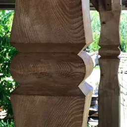 Detal rzeźbionej, drewnianej kolumny z widocznymi słojami drewna i elementami dekoracyjnymi, w tle zieleń drzew i stos drewna.