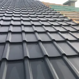 Montaż szarego pokrycia dachowego z widoczną konstrukcją drewnianą i zieloną folią w górnej części kadru, ukazujący postęp prac.