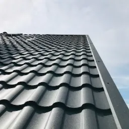 Szczegółowe ujęcie fragmentu dachu pokrytego ciemnoszarą, metalową dachówką modułową, widoczne łączenia i faktura materiału, na tle jasnego nieba.