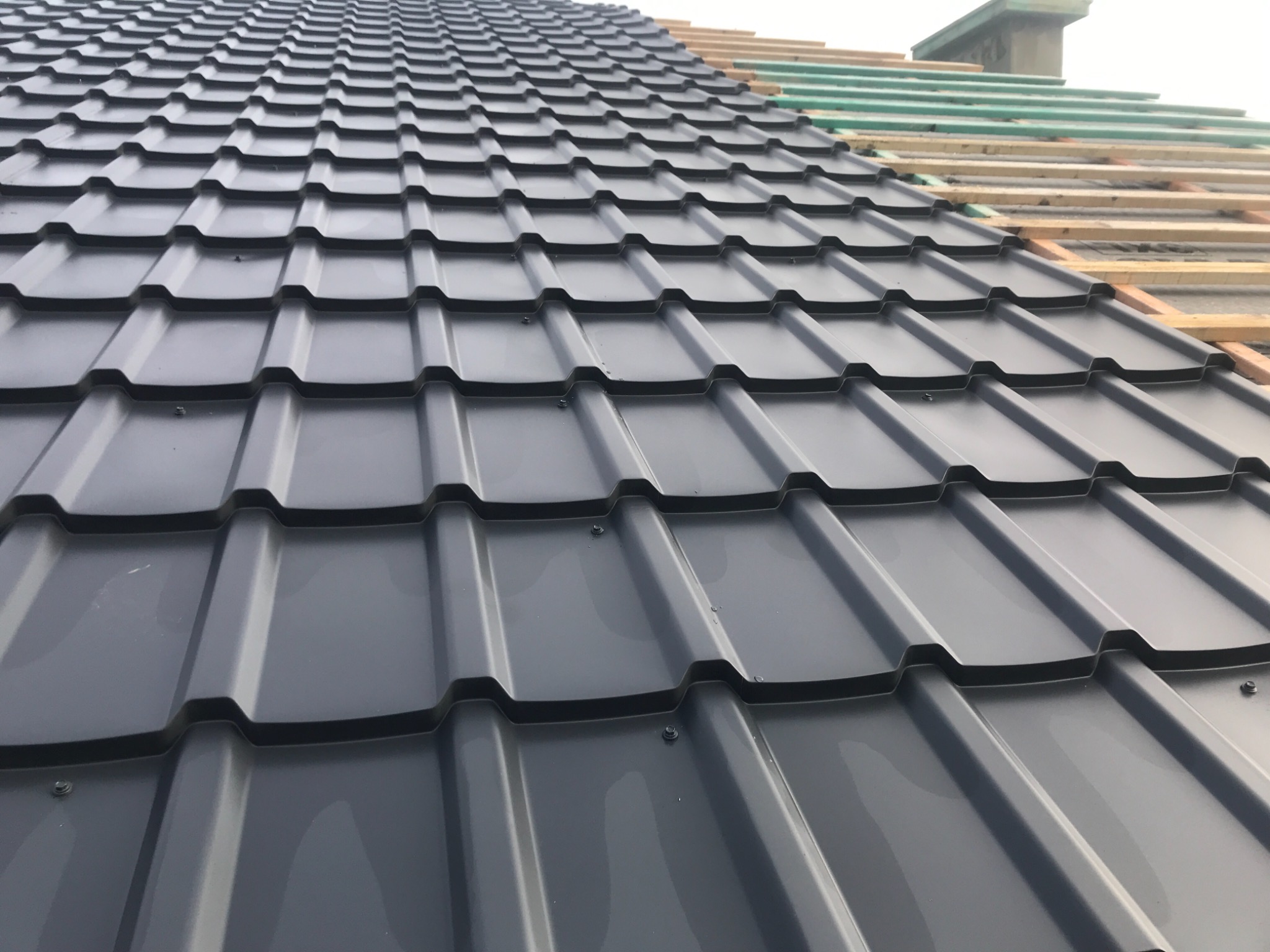 Montaż szarego pokrycia dachowego z widoczną konstrukcją drewnianą i zieloną folią w górnej części kadru, ukazujący postęp prac.