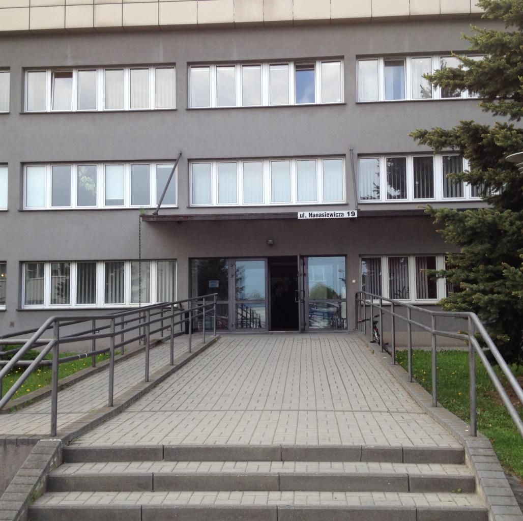 Szary budynek biurowy z podjazdem dla niepełnosprawnych i tabliczką z adresem 'ul. Hanasiewicza 19'. Widok z zewnątrz, dzień.