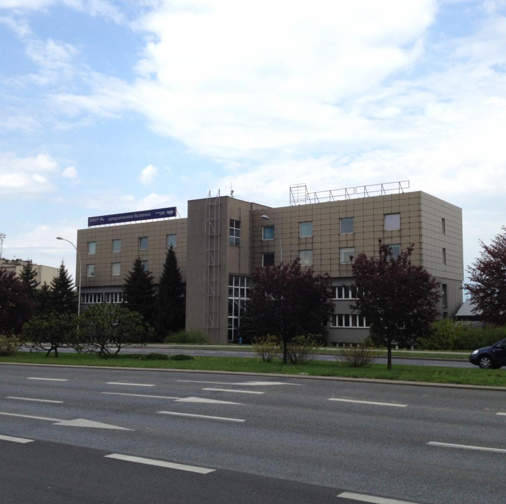 Szary budynek biurowy z napisem 'DIGIT AL oprogramowanie dla biznesu' na dachu, widoczny z ulicy z zielenią i przejeżdżającym samochodem.