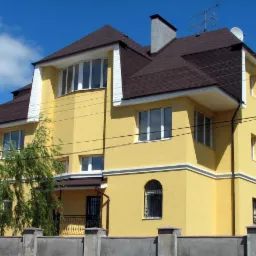 Żółty dom z brązowym dachem i białymi detalami architektonicznymi, widoczny zza betonowego ogrodzenia, w słoneczny dzień.