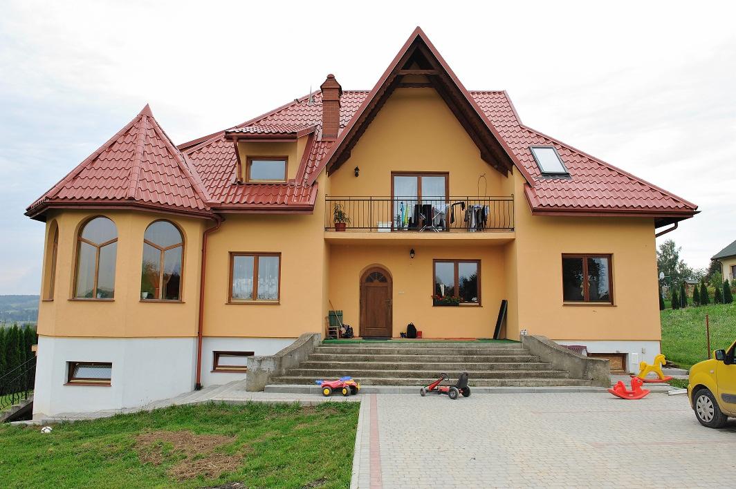 Dwupiętrowy dom z żółtą elewacją i czerwoną dachówką, widok z przodu na schody wejściowe z zabawkami dziecięcymi, balkon z suszącym się praniem.