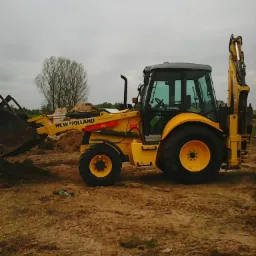 Żółta koparko-ładowarka New Holland na placu budowy, z widocznym łyżką i ramieniem kopiącym, na tle pochmurnego nieba i drzewa bez liści.
