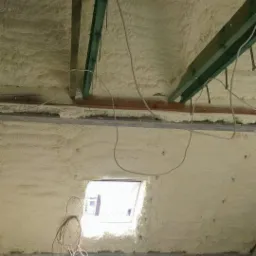 Poddasze po natrysku pianki PUR, widoczne drewniane elementy konstrukcyjne i okno dachowe. Biała pianka szczelnie wypełnia przestrzeń, izolując poddasze.