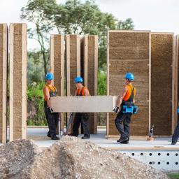 Multi Energia Sp. z o.o. - Montaż ekologicznego domu modułowego: czterech robotników w kaskach przenosi panel ściany z naturalnych materiałów na placu budowy, widoczne kolejne elementy konstrukcji.