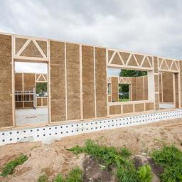 Multi Energia Sp. z o.o. - Wznoszenie ścian domu modułowego ze słomy na fundamencie z izolacją. Widoczne drewniane ramy i wypełnienie ścian słomą. Częściowo ukończona konstrukcja, widok z zewnątrz.