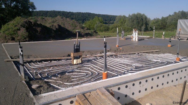 Fundament z instalacją ogrzewania podłogowego przed zalaniem betonem, widoczne białe rury w kształcie meandrów, zbrojenie i szary beton. W tle zielone wzgórze.