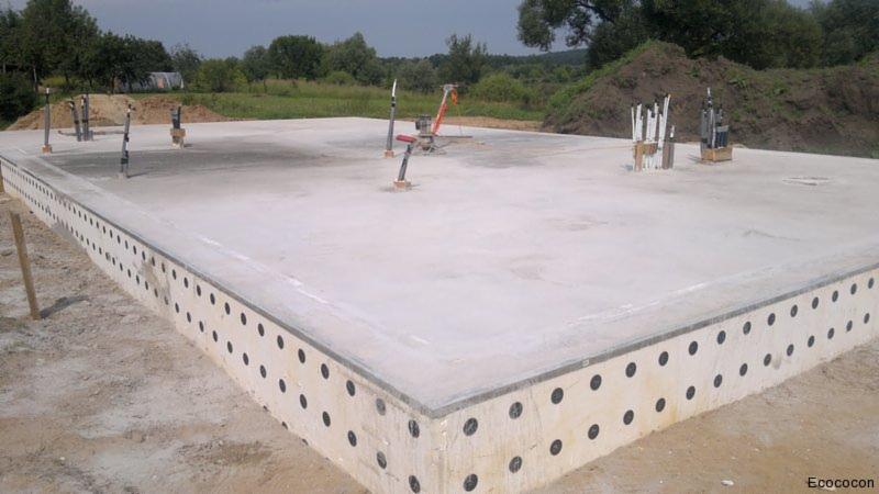 Płyta fundamentowa z izolacją obwodową, widoczne wystające elementy instalacji. Tło: zielona roślinność. Realizacja z użyciem szalunków traconych.