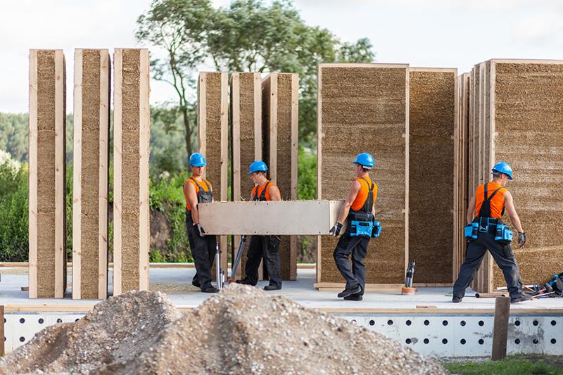 Montaż ekologicznego domu modułowego: czterech robotników w kaskach przenosi panel ściany z naturalnych materiałów na placu budowy, widoczne kolejne elementy konstrukcji.