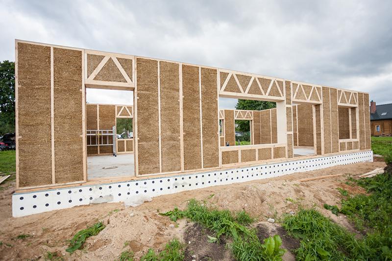 Wznoszenie ścian domu modułowego ze słomy na fundamencie z izolacją. Widoczne drewniane ramy i wypełnienie ścian słomą. Częściowo ukończona konstrukcja, widok z zewnątrz.