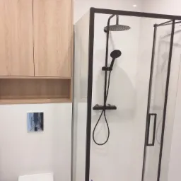 Nowoczesna łazienka z kabiną prysznicową typu walk-in z czarnymi profilami, wiszącą szafką z jasnego drewna i podtynkową toaletą.